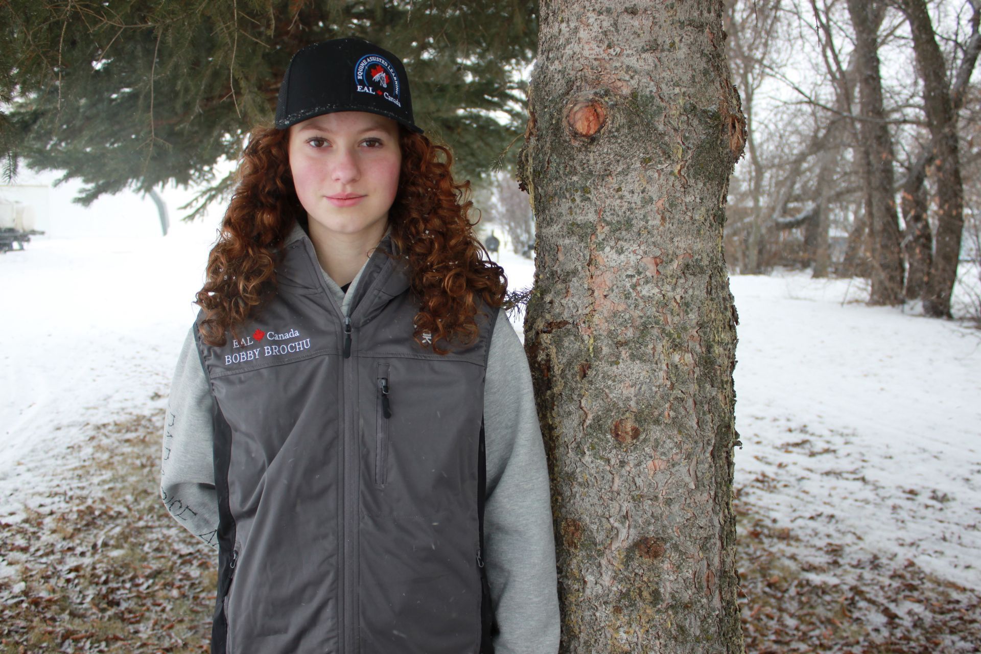 Woman in black hat and hoodie smiles next to a tree in a snowy setting.