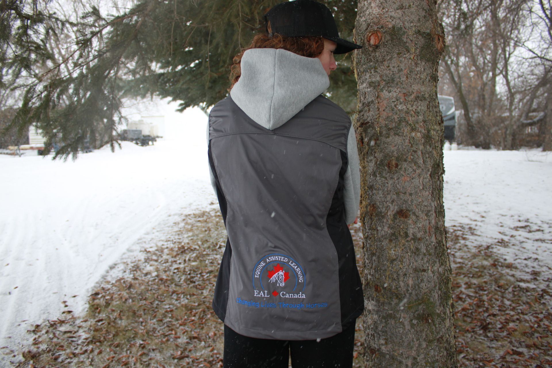 Person with curly hair wearing a vest and hat, standing near a tree in a snowy environment.