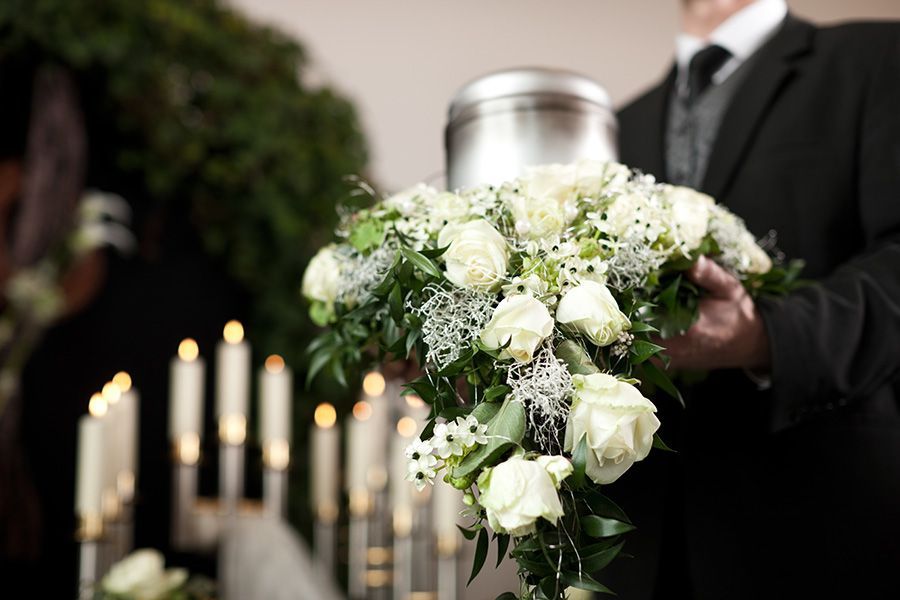 A man in a suit is holding a bouquet of white roses and a urn at a funeral.