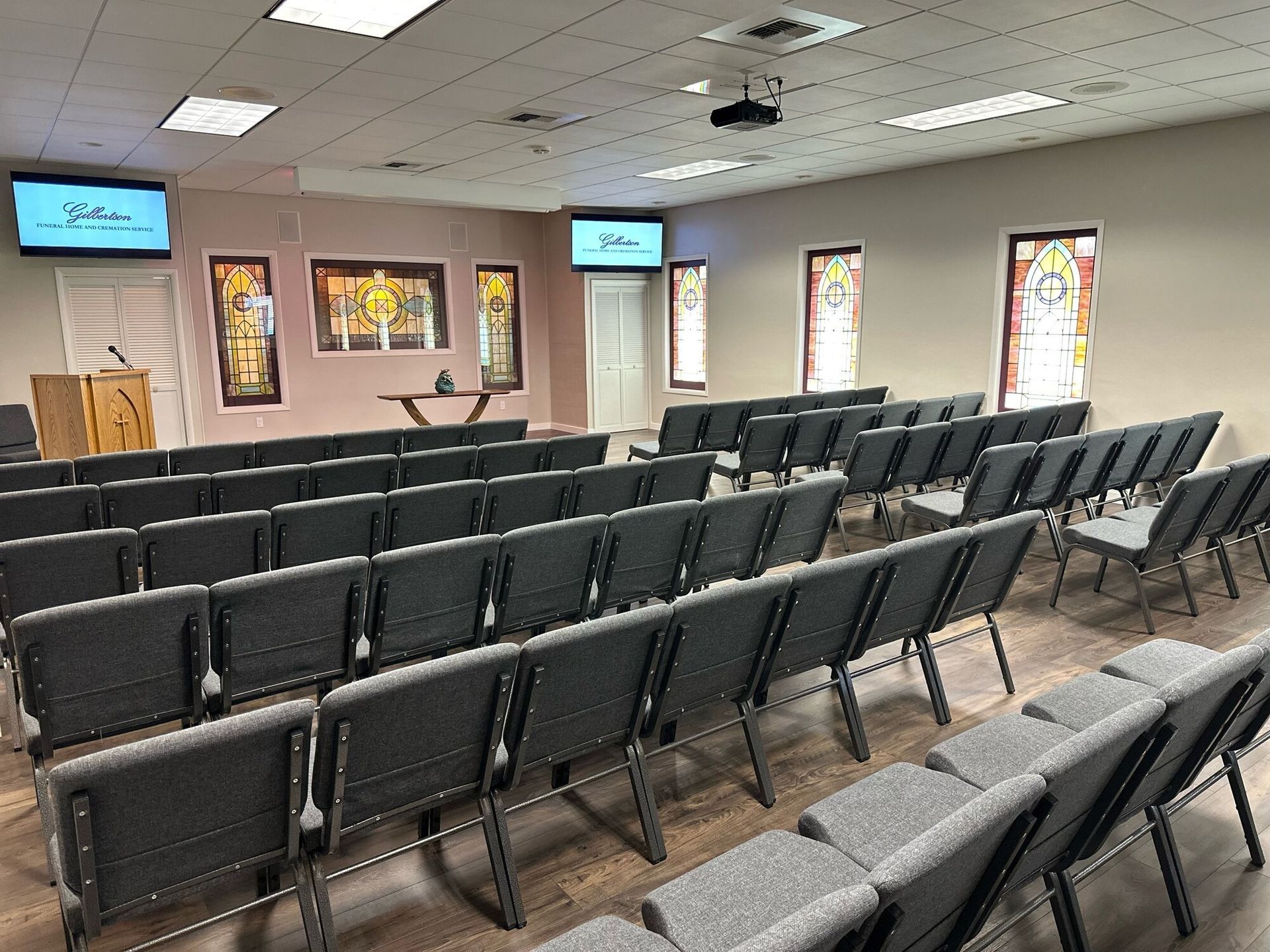 Empty church sanctuary with rows of gray chairs, screens, and stained-glass windows.