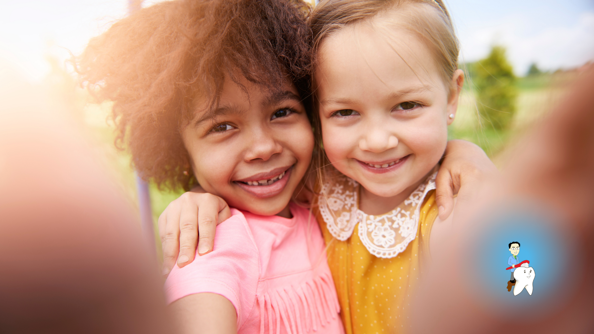 Two young girls of different races smiling, arms around each other, taking a selfie outdoors.