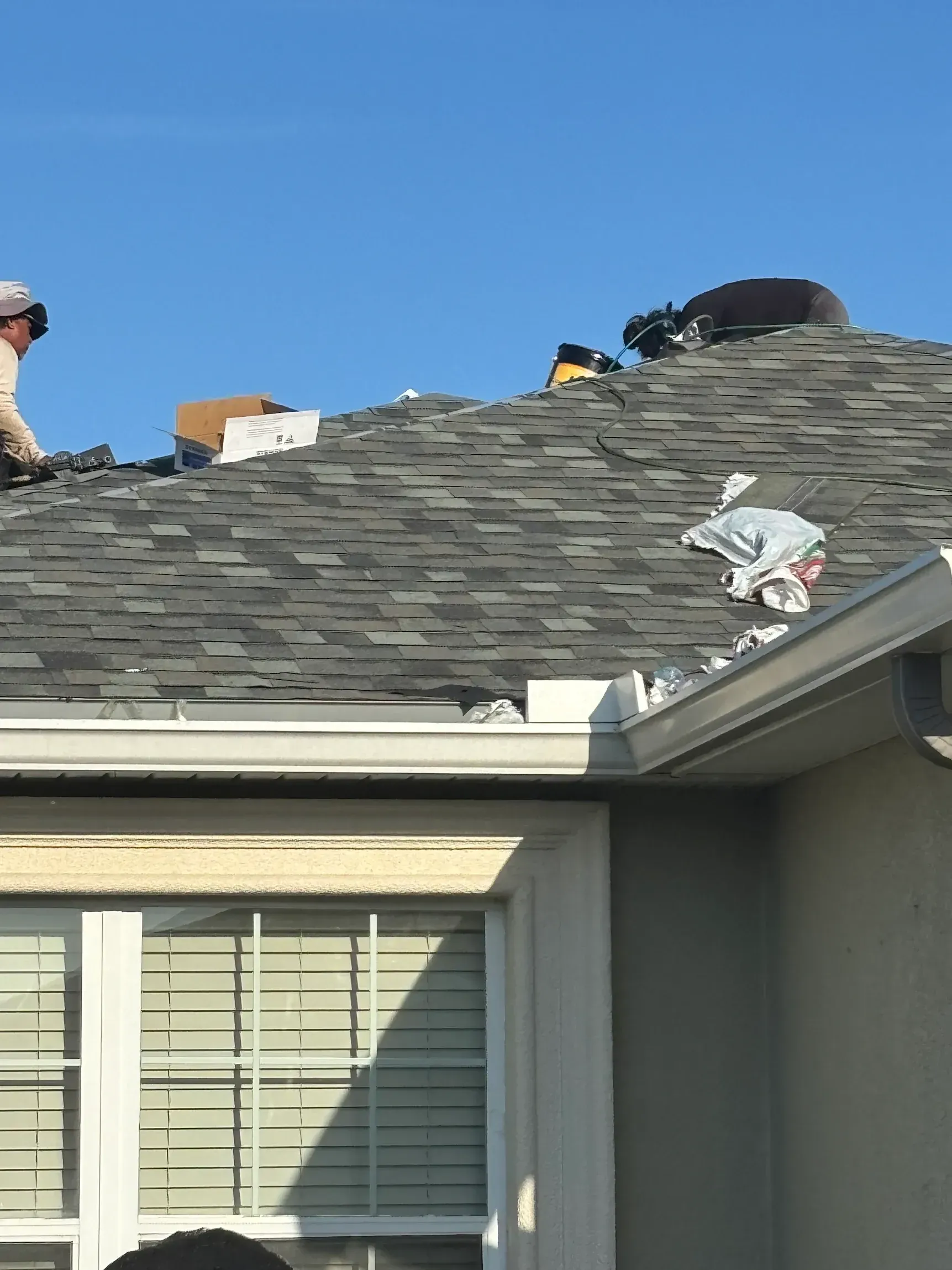 Roofers working on a gray shingled house roof under a clear blue sky