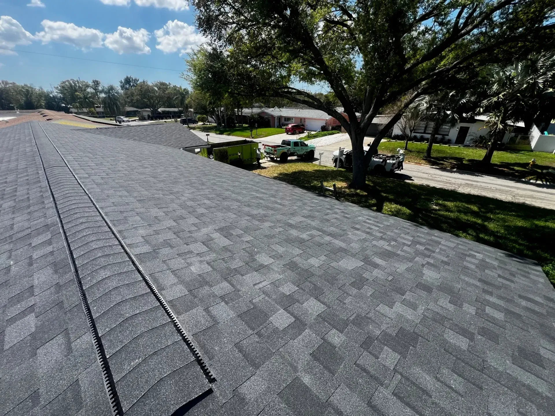 Shingled rooftop beside a tree-lined street under a bright blue sky with clouds
