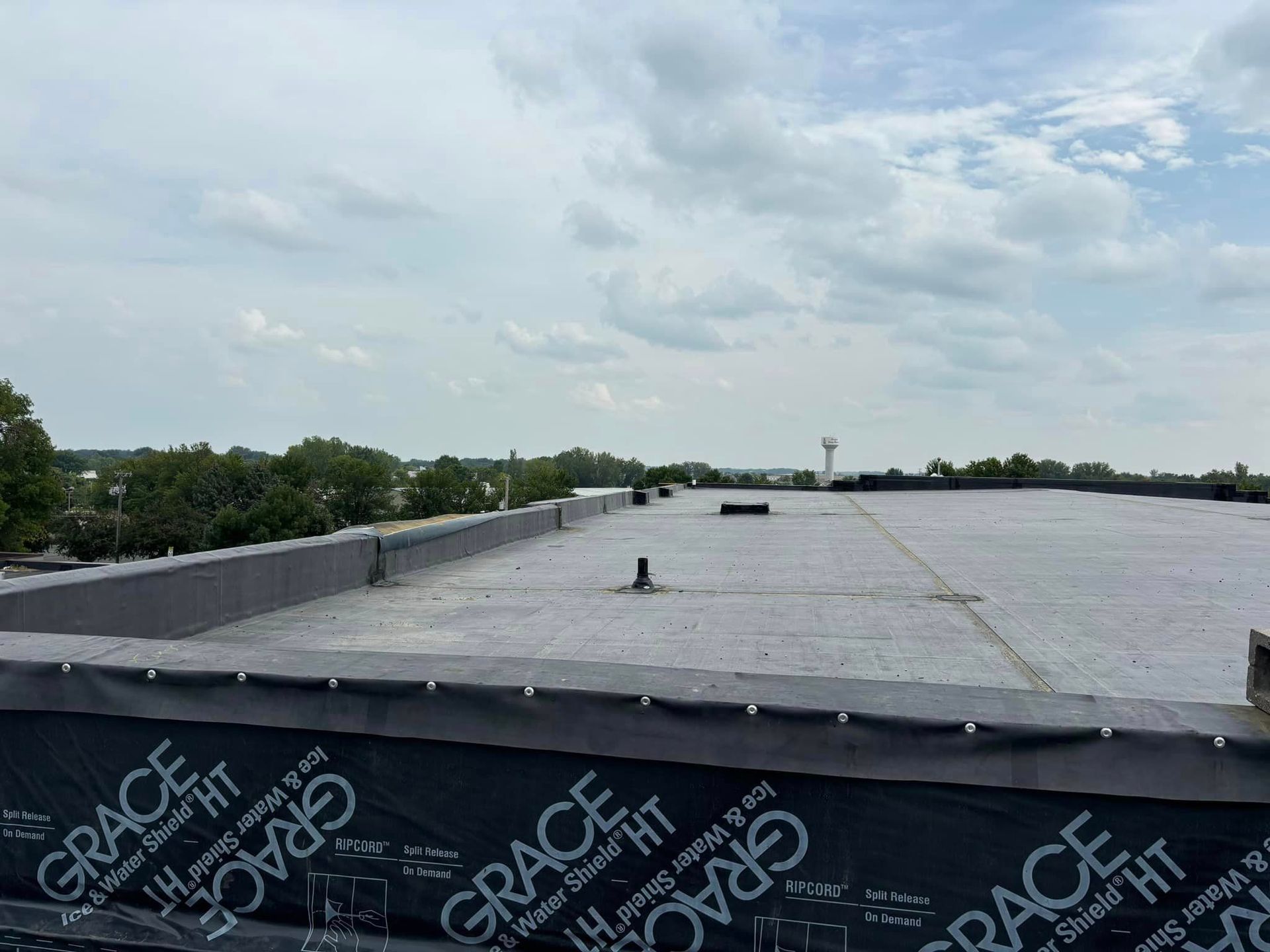 Flat rooftop under a cloudy sky, with black roofing material and trees in the distance