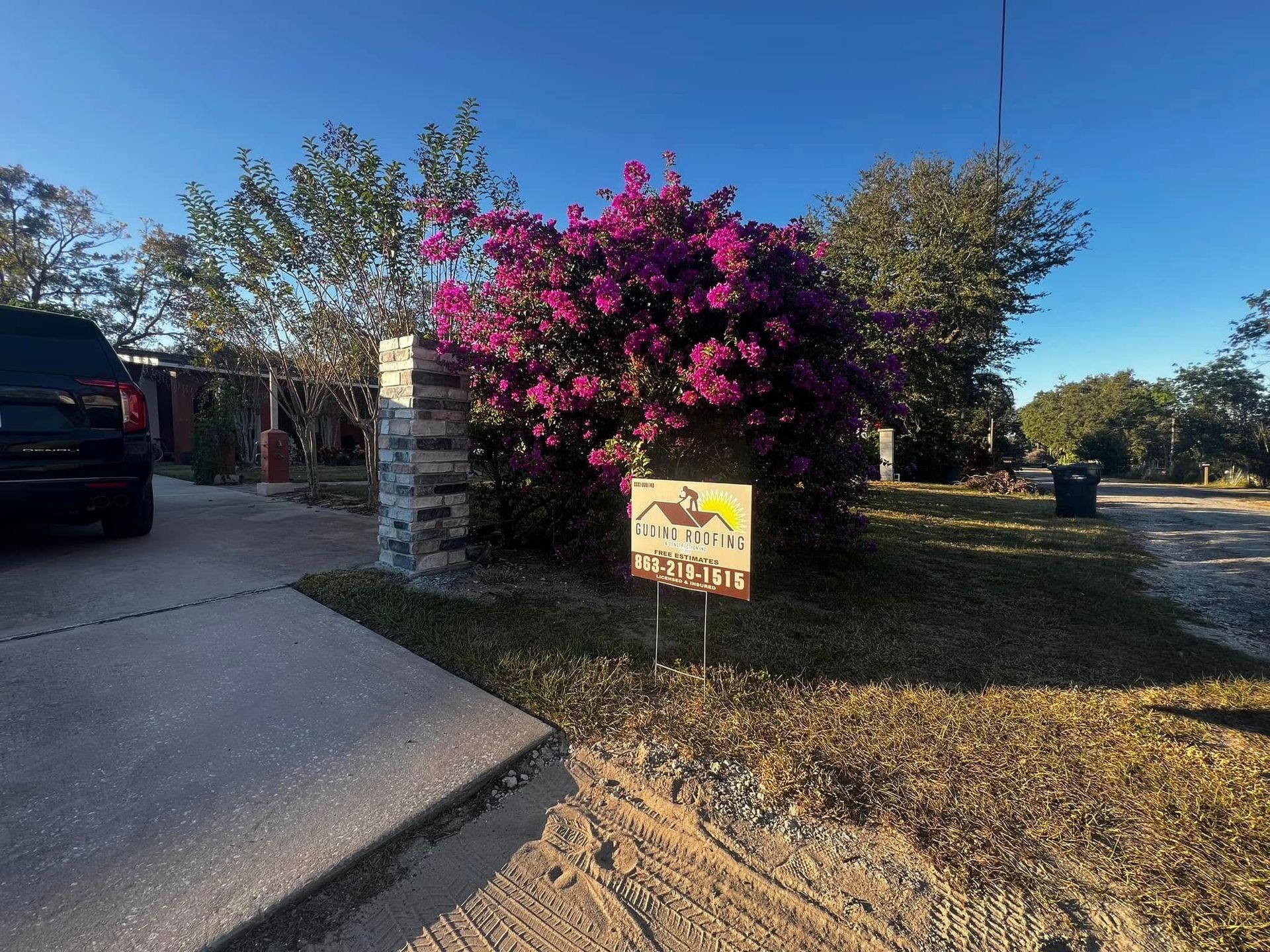 Bright pink flowering bush by a driveway with a yard sign in front under a clear blue sky