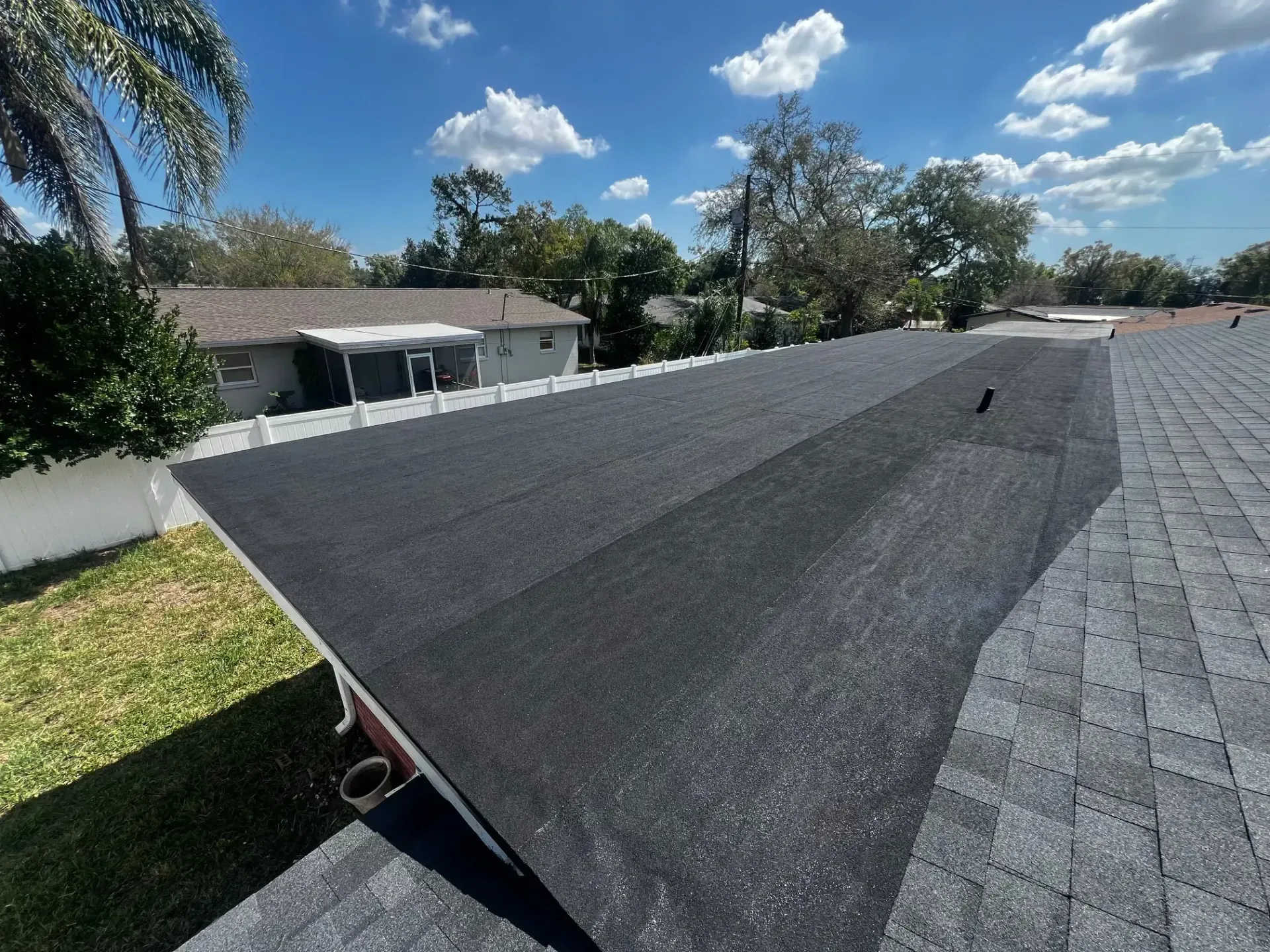 Roof being reroofed with dark shingles under a sunny blue sky in a suburban neighborhood