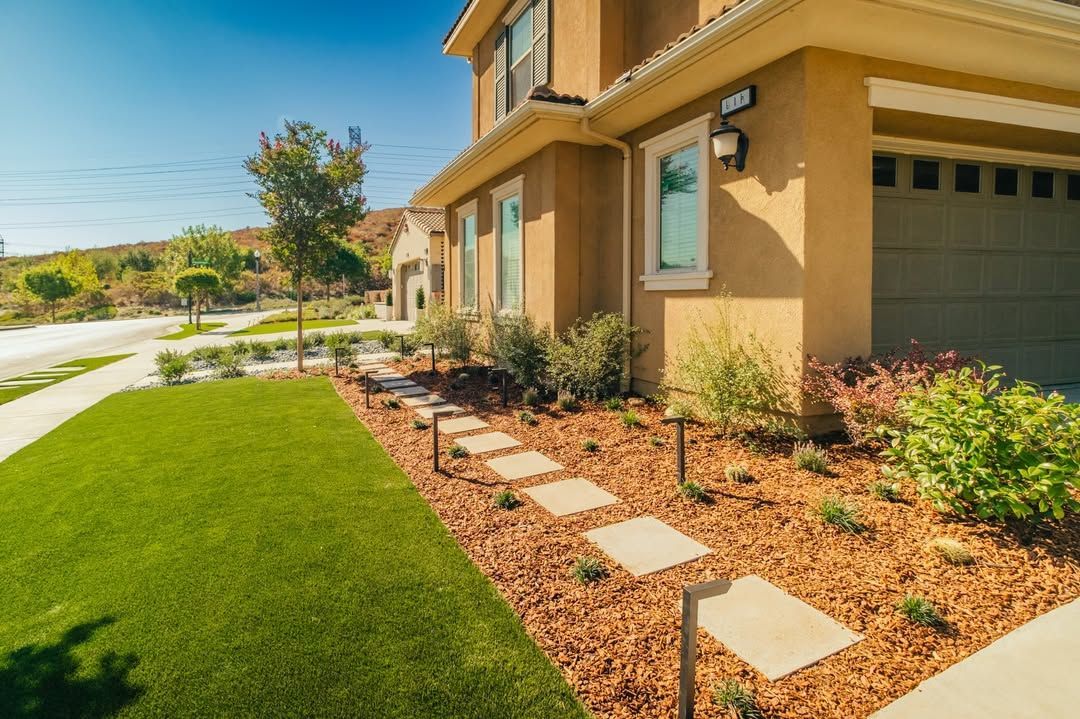 A two-story house with a green lawn, stepping stone path, and a brown mulch-covered bed.