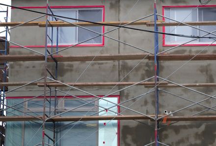 Picture of a stucco houses exterior with scaffolding setup in front of it. The house has its windows taped for protection and has a new stucco base coat.