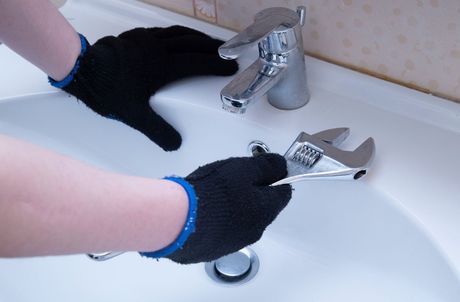 Picture of a handyman plumber wearing black gloves holding a wrench to a bathroom faucet in preparation of repair.