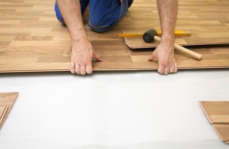 Picture of a handyman installing laminate floor. The floor installer is on his hand and knees' inserting a plank wearing blue pants.