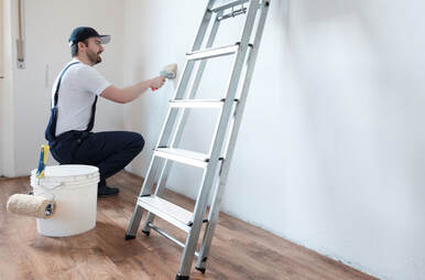 Handyman painter picture of a worker in blue overalls and white T-shirt painting a wall blue with a roller. The worker is kneeled down crouched beside his painting tools and step ladder.