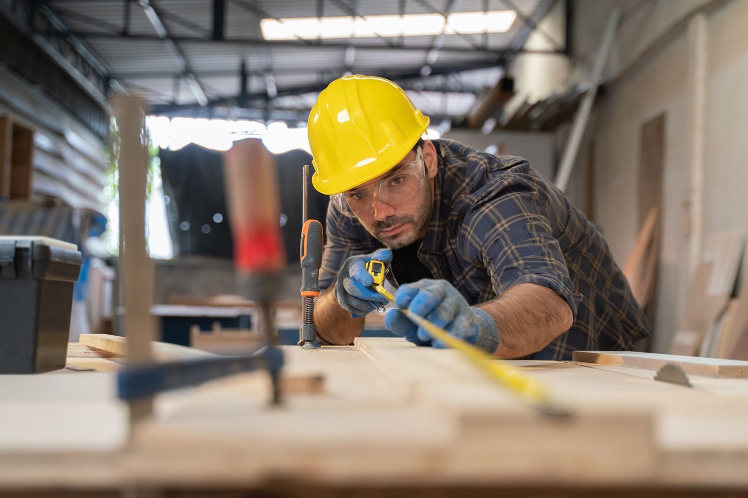 A man is measuring a piece of wood with a tape measure in a workshop.