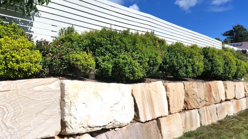 A Stone Wall Surrounded by Bushes and a White Fence — Brendan's Landscaping and Fencing In Bundaberg, QLD