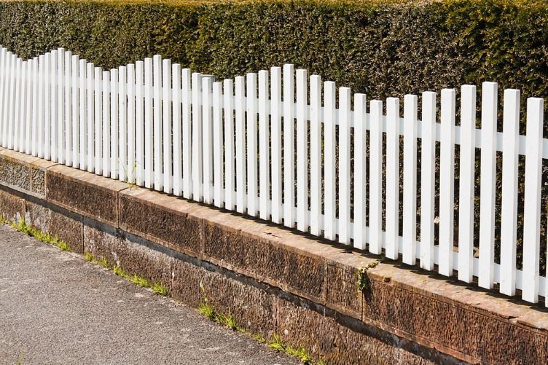 A White Picket Fence is Sitting Next to a Stone Wall — Brendan's Landscaping and Fencing In Elliot Heads, QLD