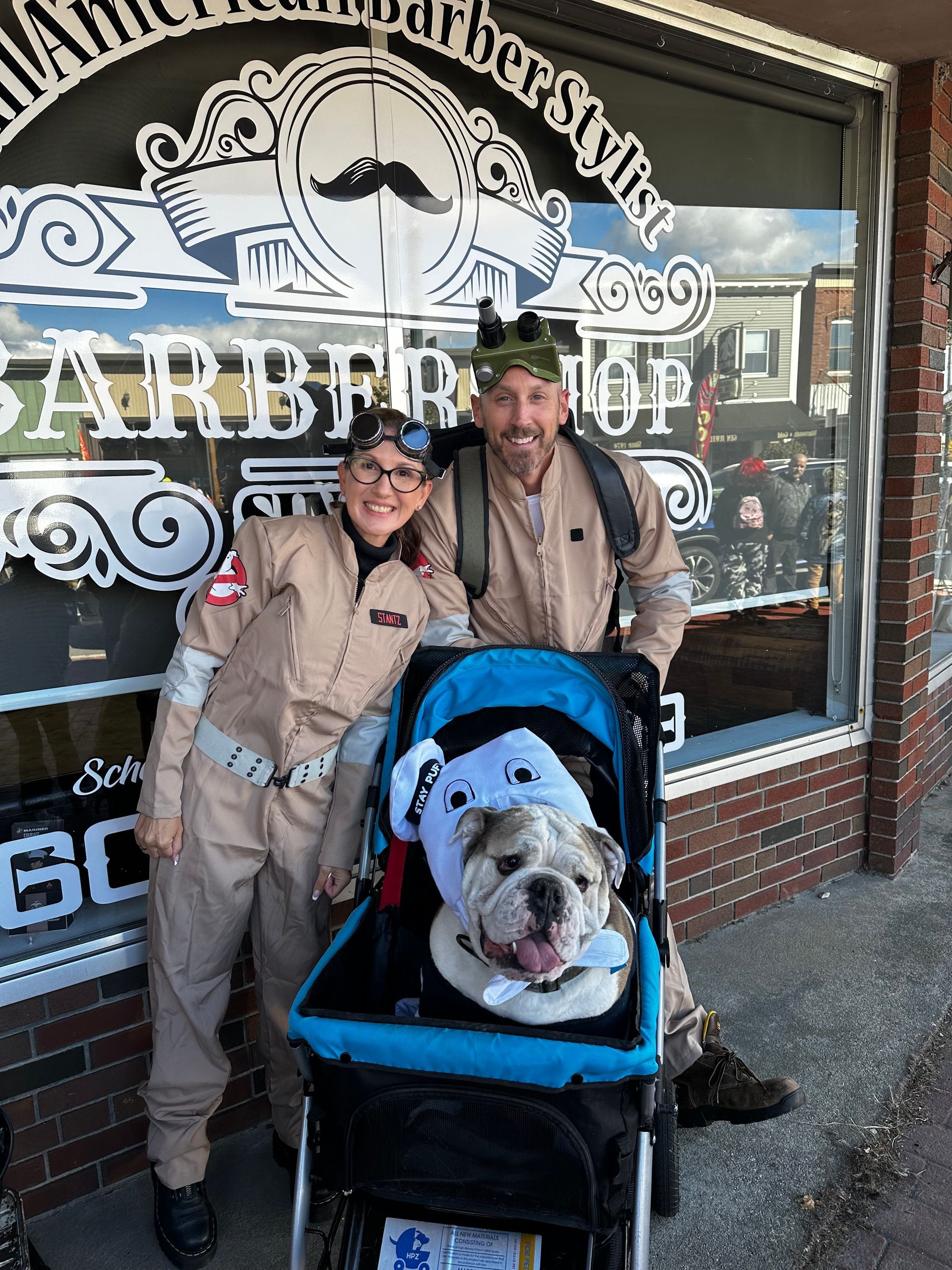 Two people in Ghostbuster jumpsuits with a dog in a stroller outside a barbershop.