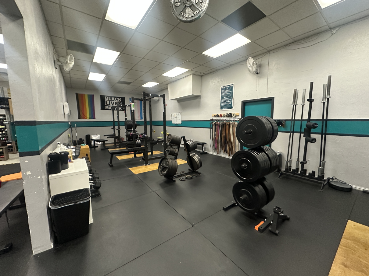 A gym interior with weight racks, barbell storage, a weight tree, and black matted flooring under white drop ceilings.