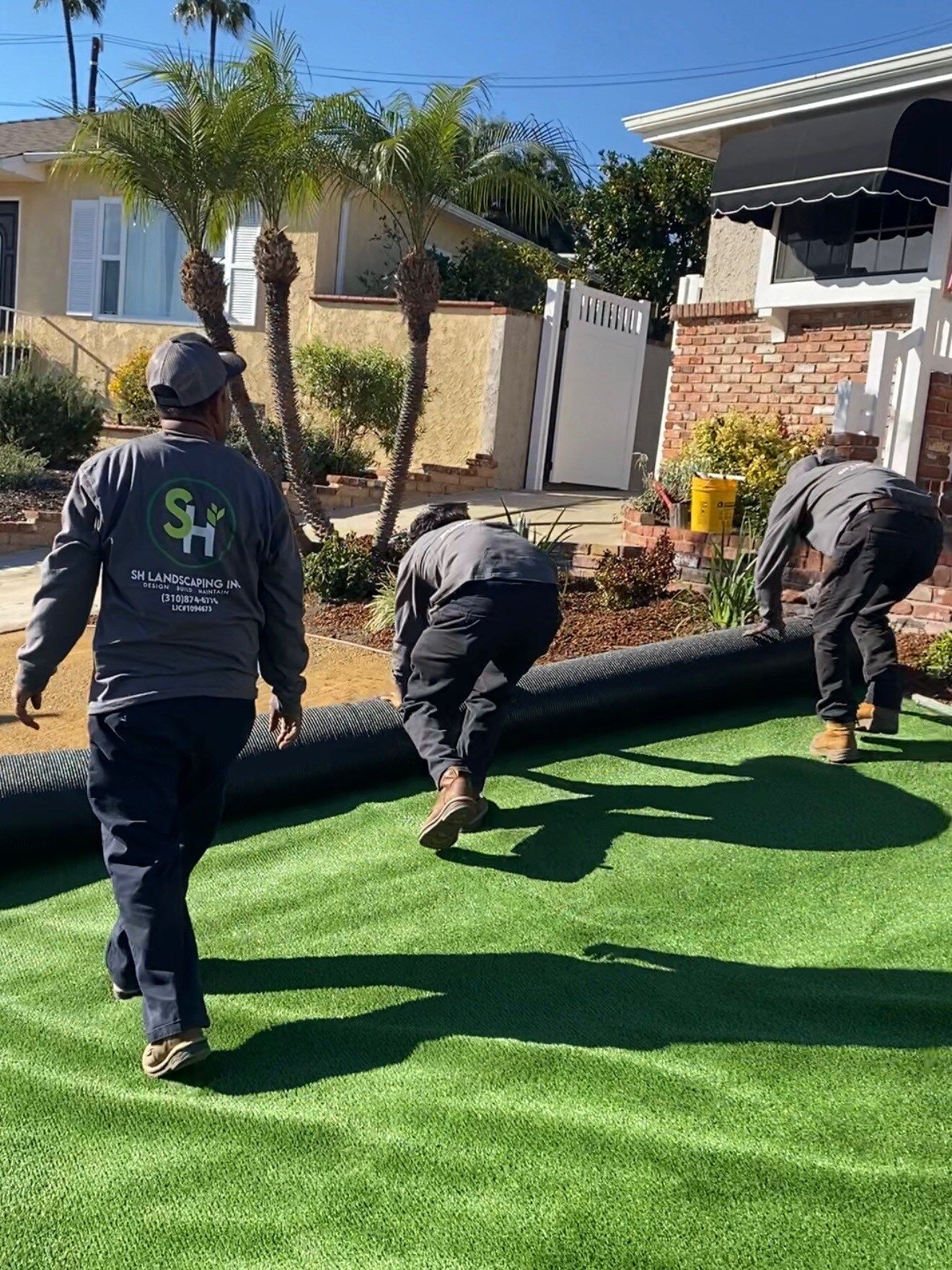 Three workers installing artificial turf on a sunny day. Green grass, gray shirts, and a suburban yard.