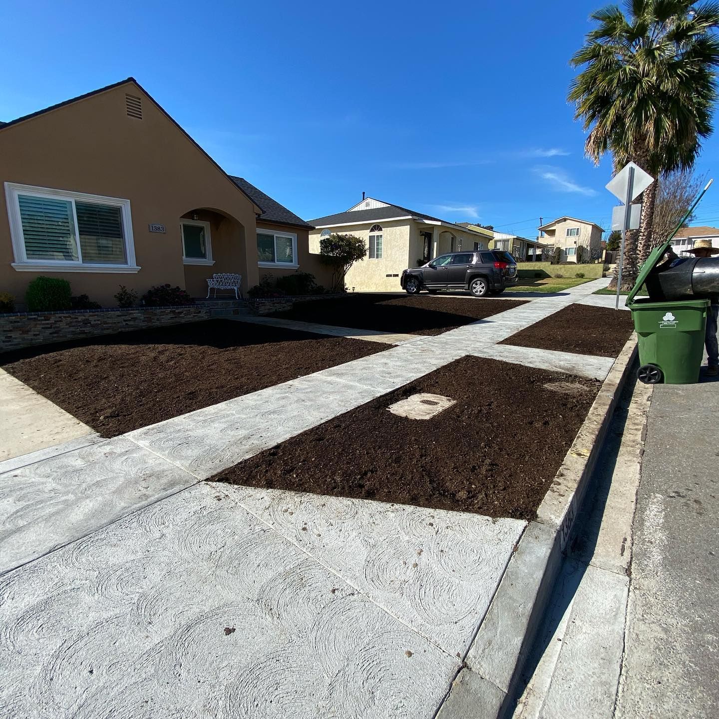House with a tan exterior, brown mulch beds, and a concrete sidewalk under a blue sky.