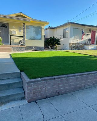 A residential lawn of bright green grass behind a brick wall, next to a sidewalk and two houses.