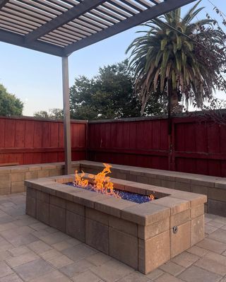 Fire pit with blue glass and flames on a patio, under a pergola, with a red fence and palm tree.