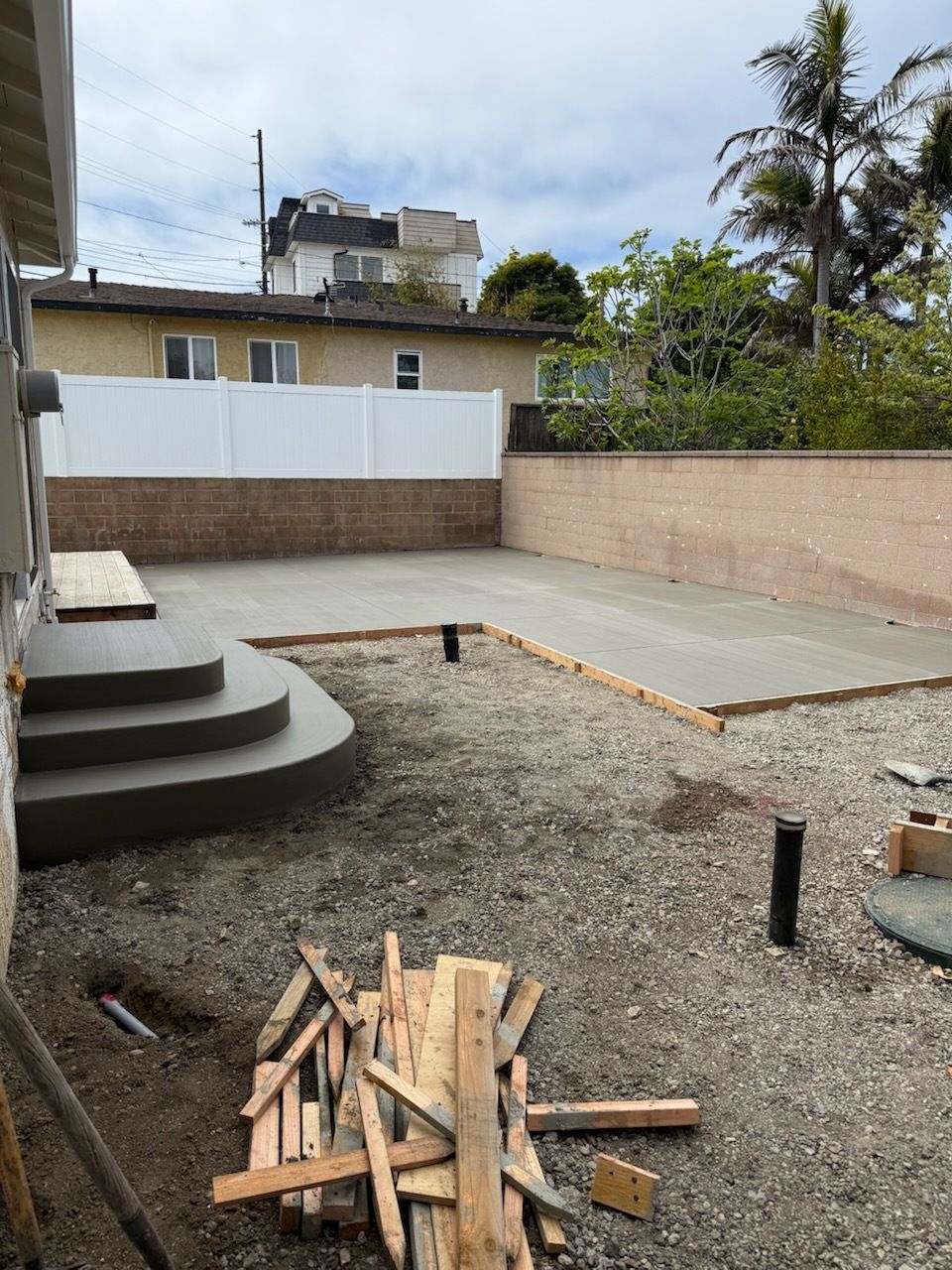 Construction site with newly poured concrete patio and stairs, gravel ground, white fence, and brick wall.