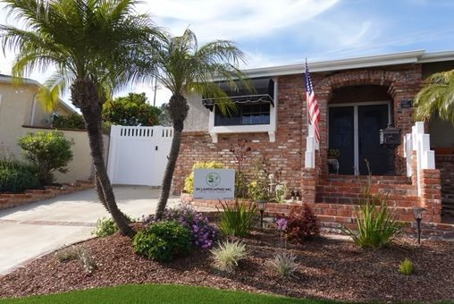 A brick home with palm trees, an American flag, and a sign in front.