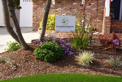 Landscaped garden bed with various plants, mulched with brown chips; brick house backdrop.