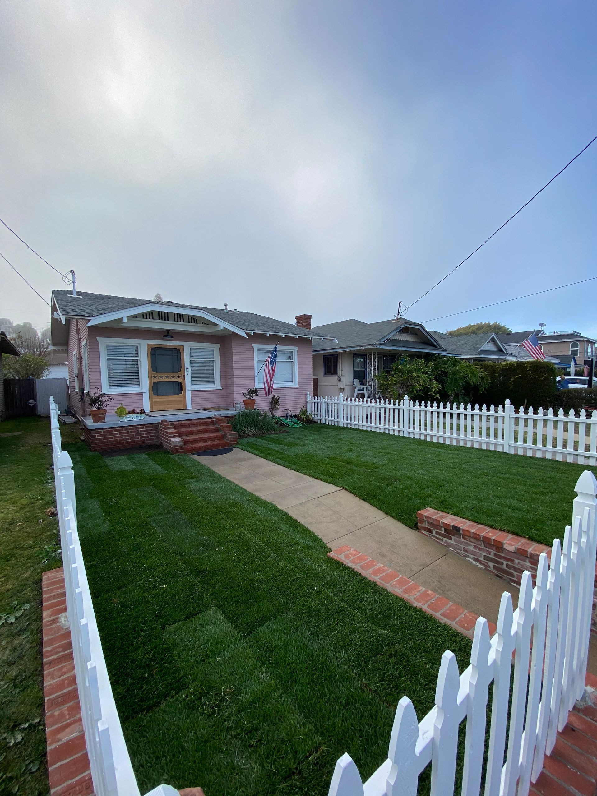 Pink house with picket fence and green lawn, under cloudy sky.