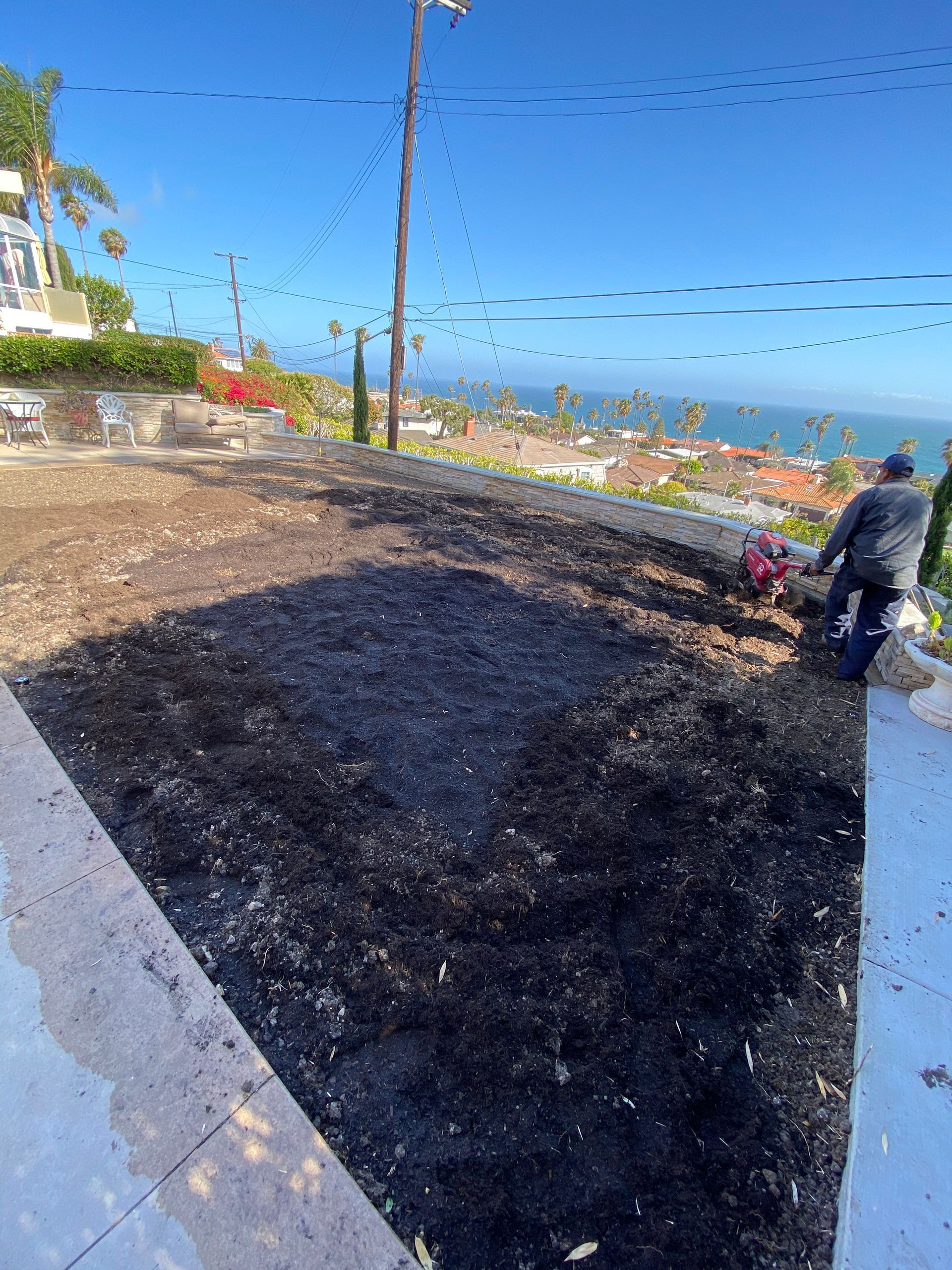 Man tilling soil on a hillside with a view of the ocean. Blue sky.