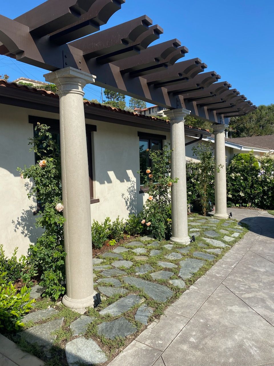 Pergola with brown beams and stone columns, with greenery and stone path.