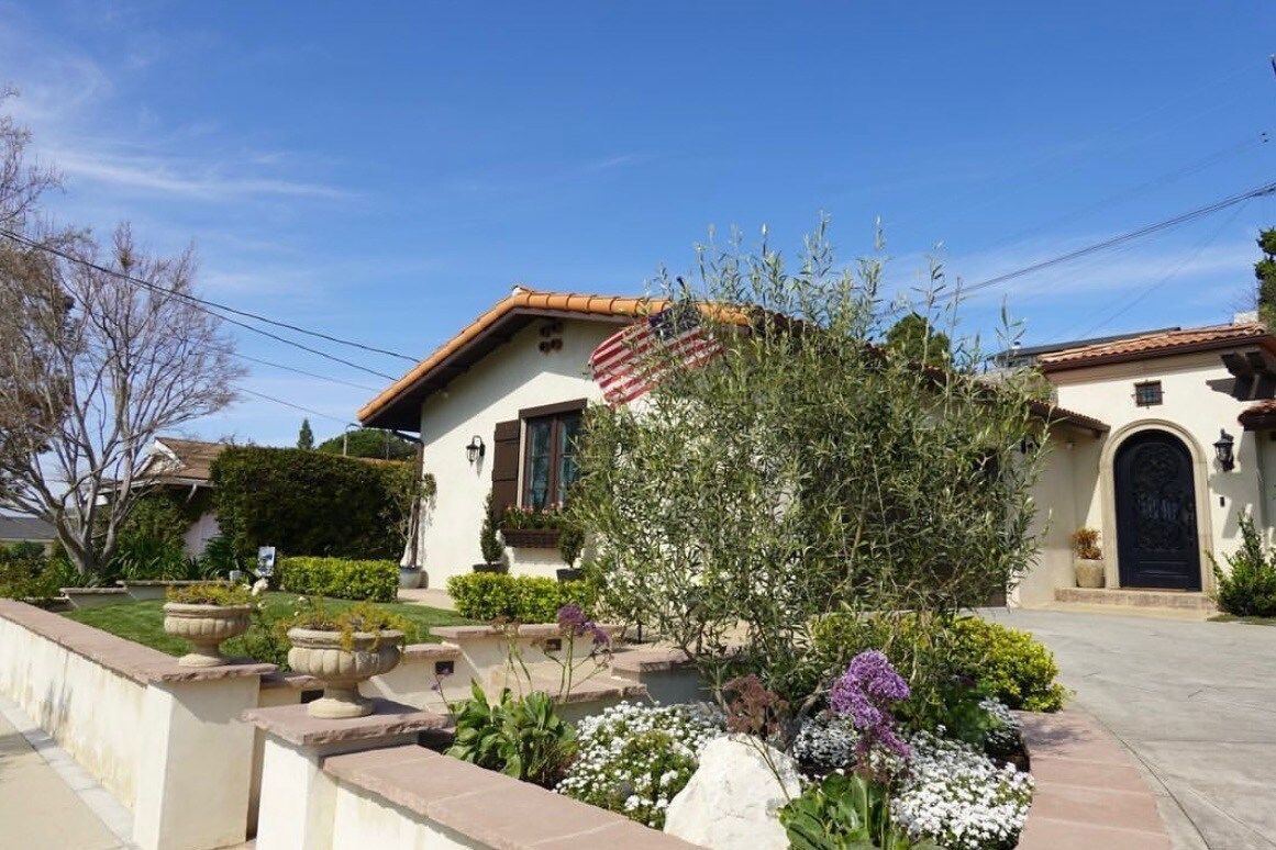 House with terracotta roof, black front door, flowering shrubs, and a blue sky.