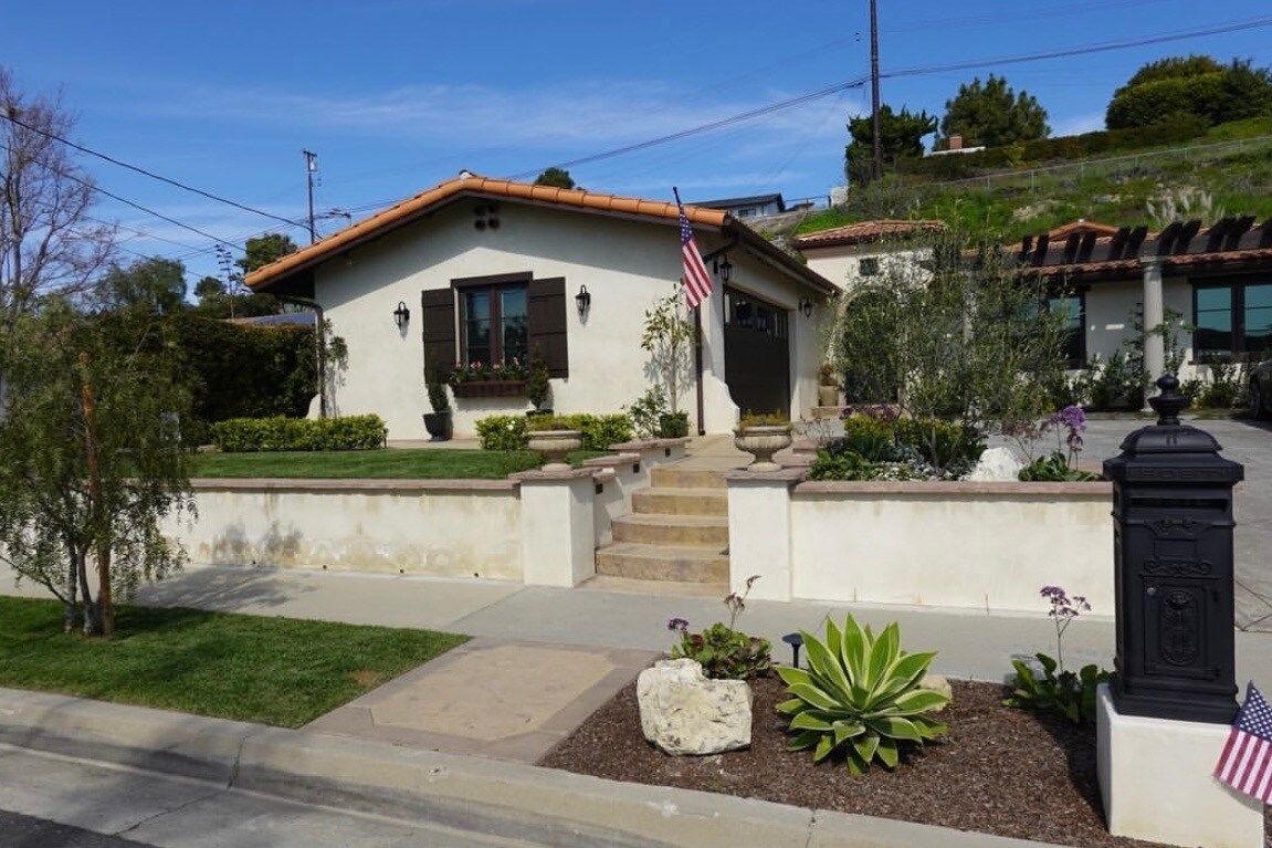 Cottage with stucco walls, stairs, and American flag. Landscaping in front, blue sky in the background.