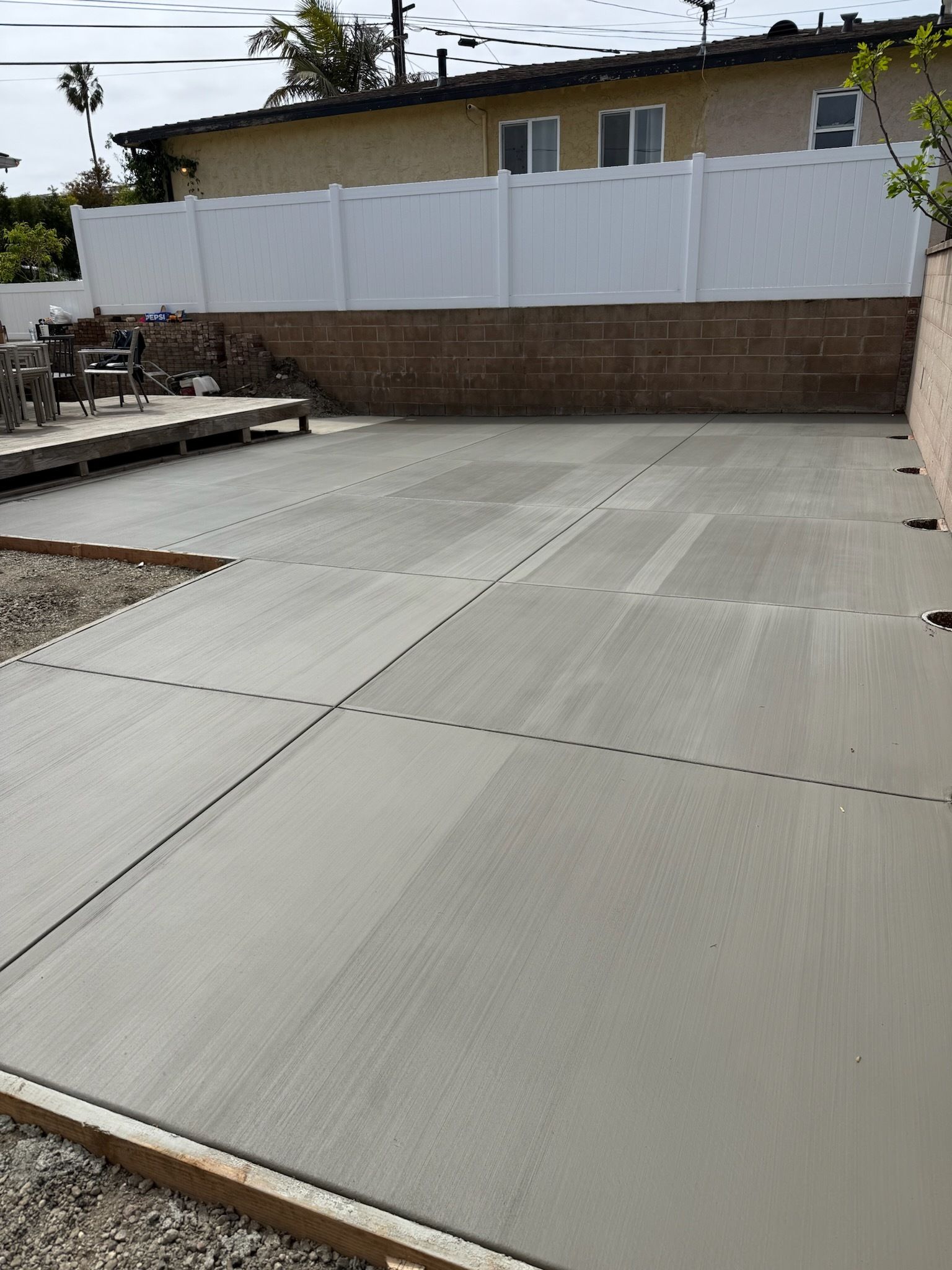 Newly poured concrete patio with expansion joints, bordered by wood, against a brick wall and white fence.
