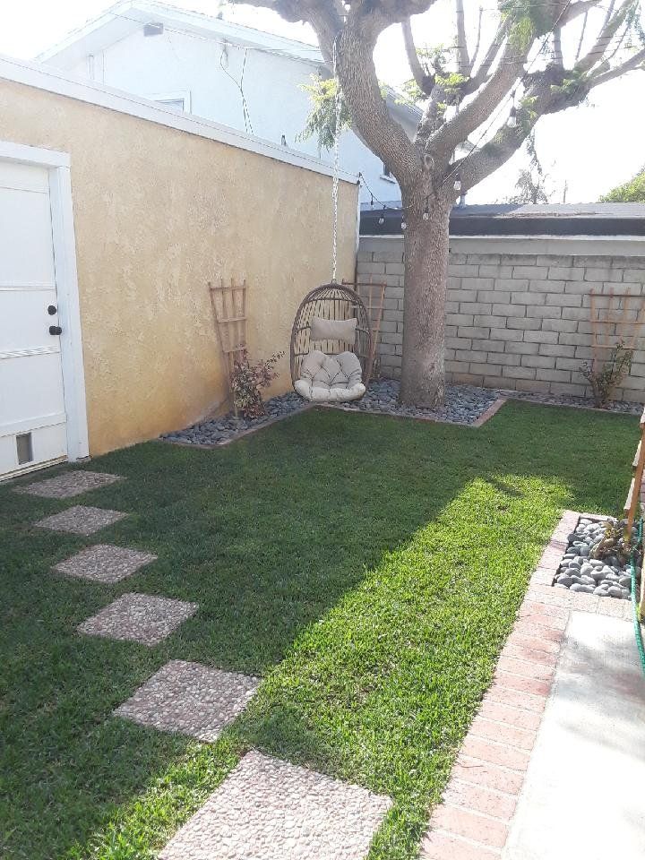 Backyard with stone path, green grass, a hanging chair near a tree, and a brick border.