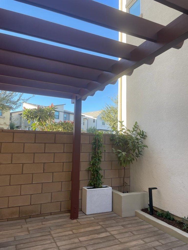 Patio with wooden pergola, brick wall, and potted plants. Sunny outdoor setting.