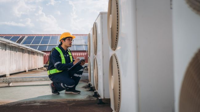 A man in a hard hat is kneeling down next to a row of air conditioners.