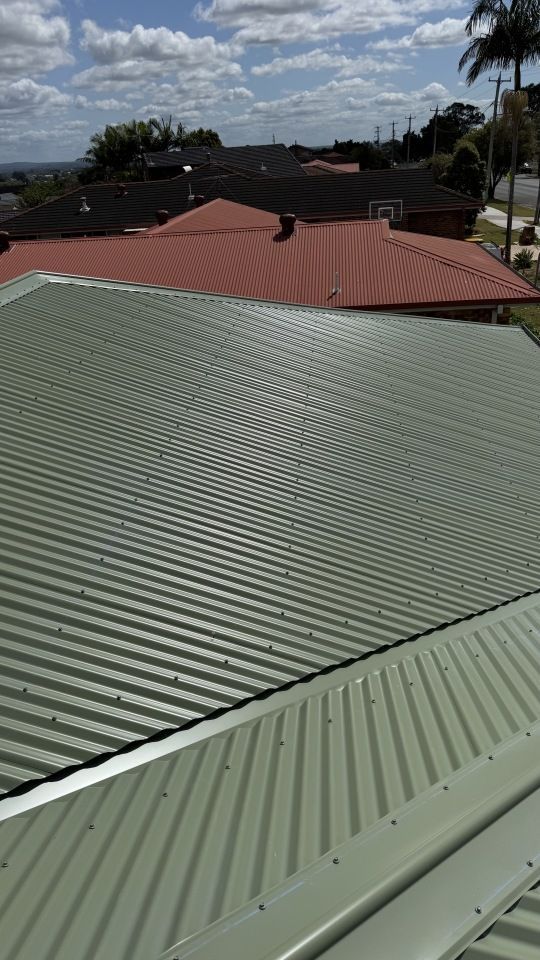 An elevated view of a green corrugated metal roof, with red and black roofs visible in the background under a blue sky.