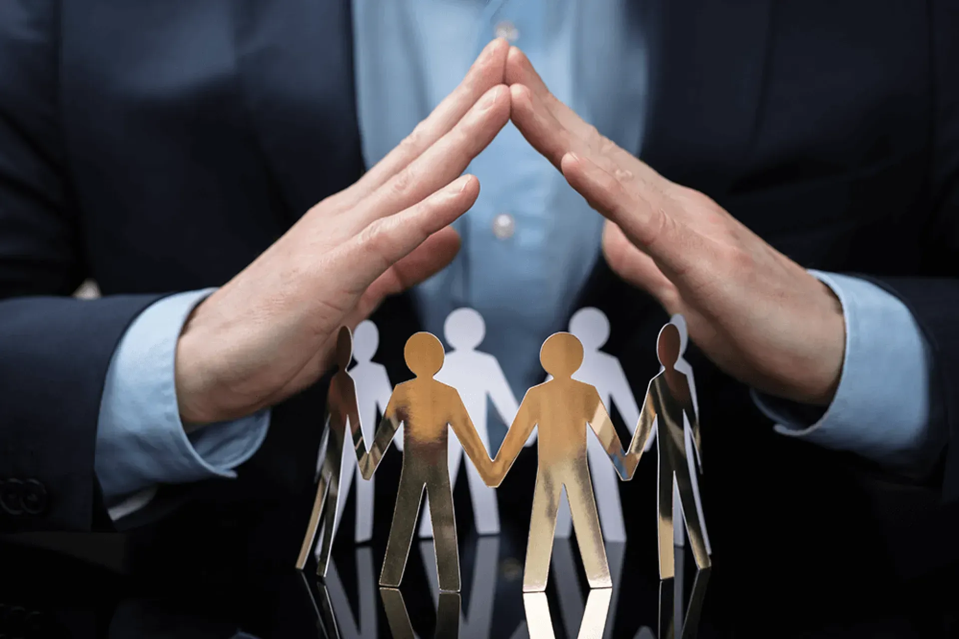 Hands forming a protective roof over paper figures in a circle, symbolizing support or protection.