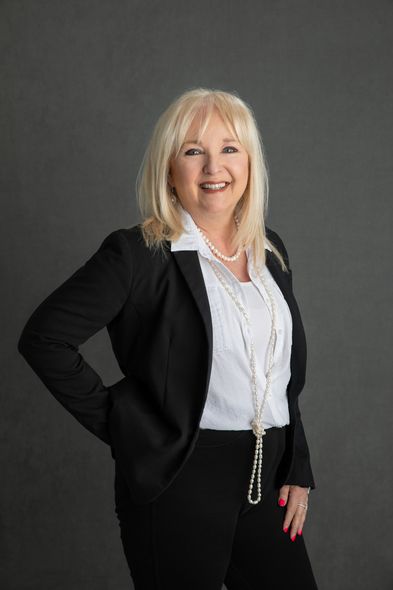 Woman in black suit smiles, arms at her side, against a gray background.