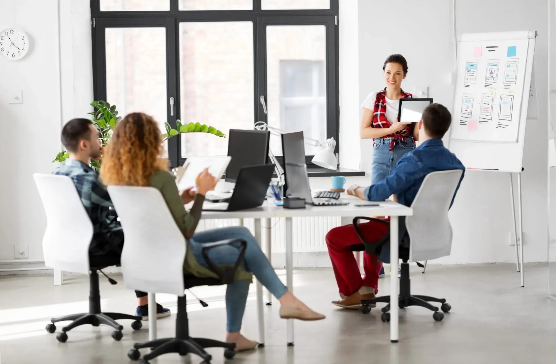 Four people in a modern office, one presenting from a tablet. A whiteboard with diagrams is in the background.