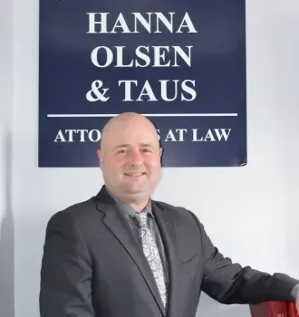 Man in suit stands in front of a law firm sign: Hanna Olsen & Taus, Attorneys at Law.