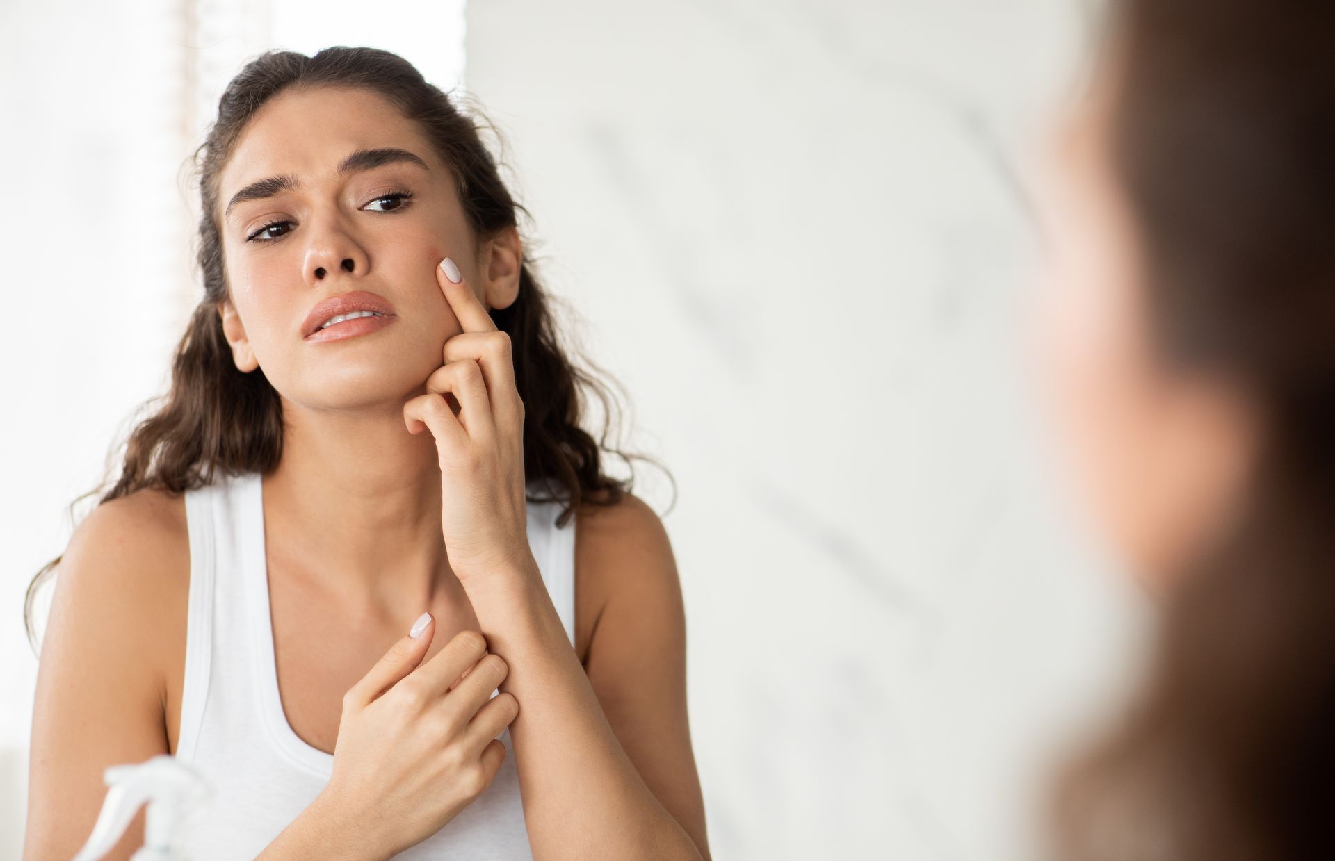 Woman looks at her face in the mirror, touching her cheek with a concerned expression. White tank top, marble background.