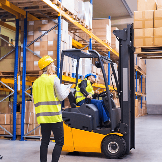 A worker in a yellow safety vest directs a forklift operator lifting a pallet in a warehouse.