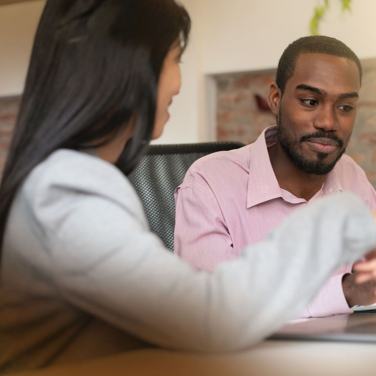 Two people in an office setting sit at a desk, appearing to have a professional conversation.
