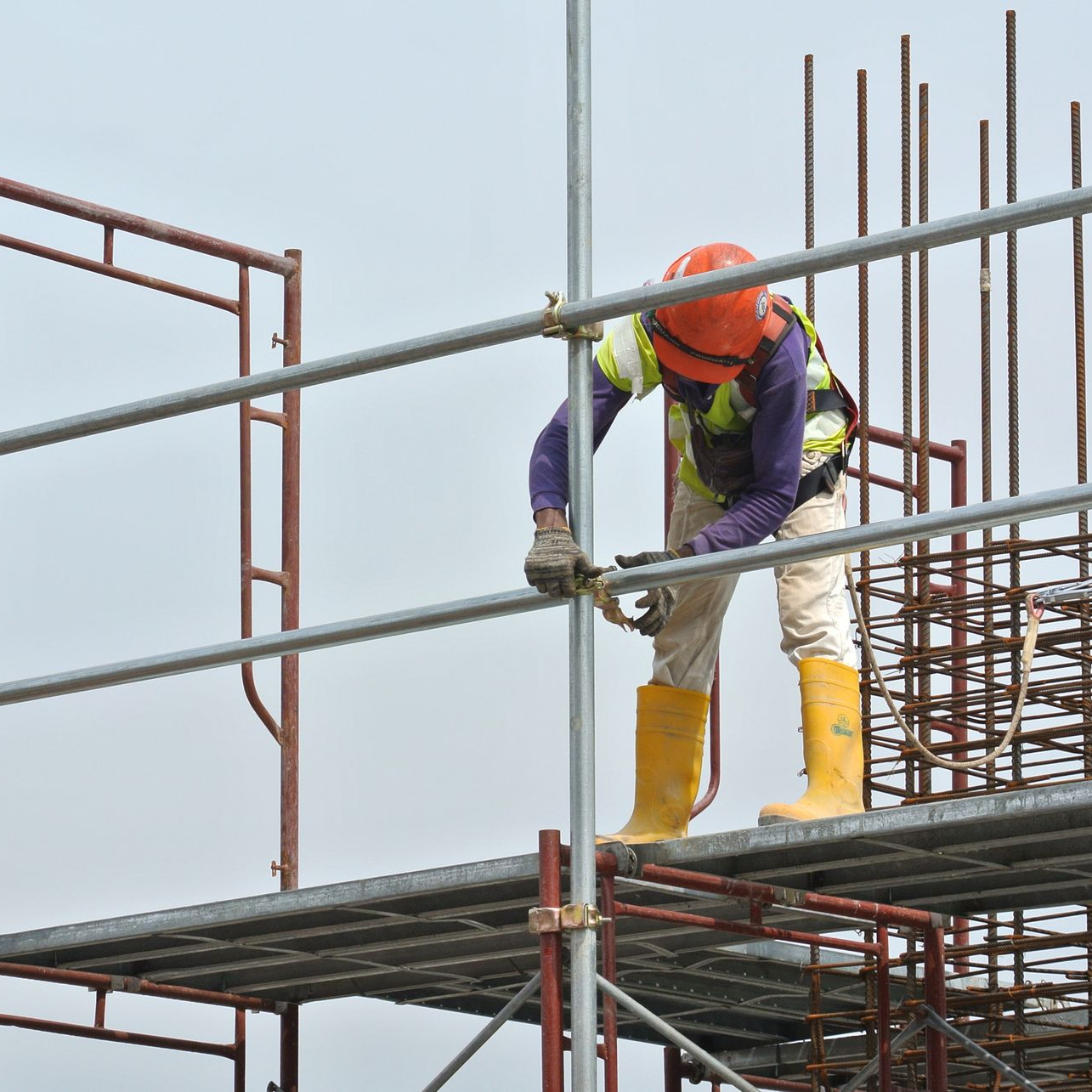 A construction worker in a hard hat, high-visibility vest, and yellow boots works on a metal scaffolding structure.