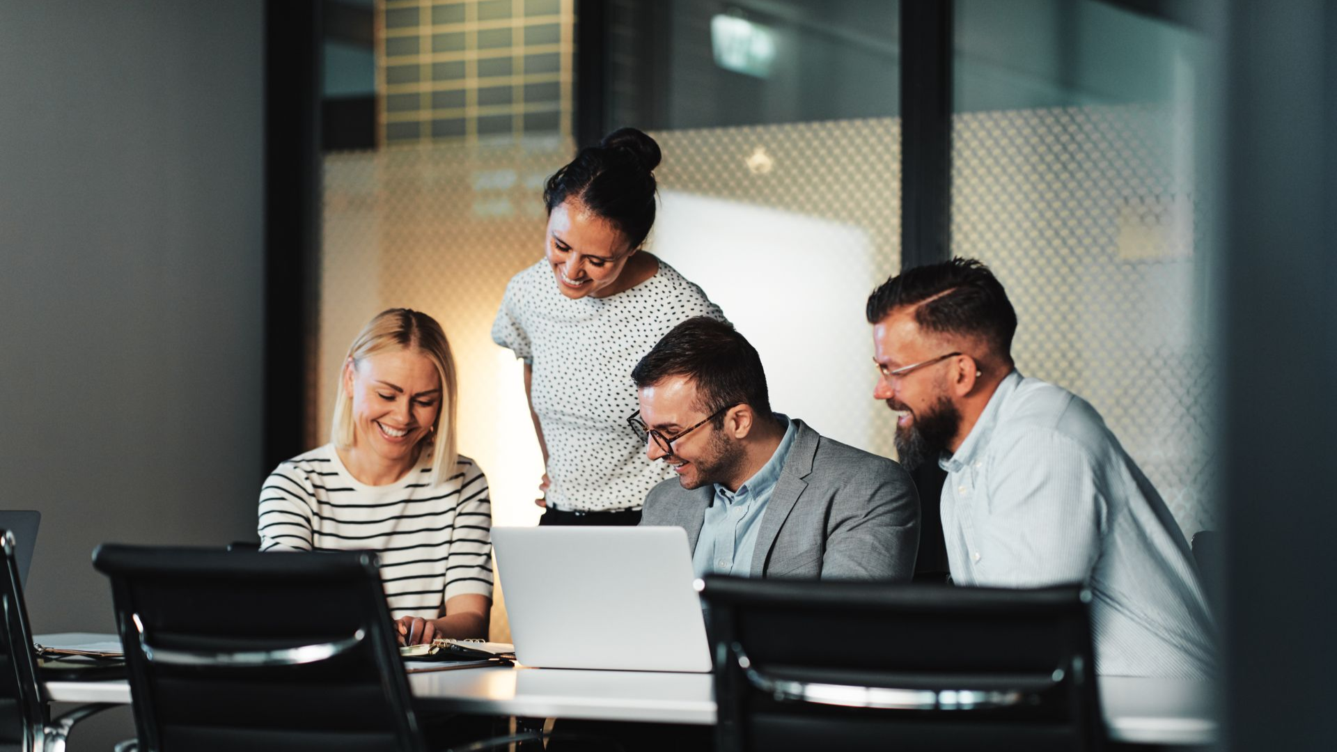 Four people collaborate around a laptop in a modern office, smiling and looking at the screen.
