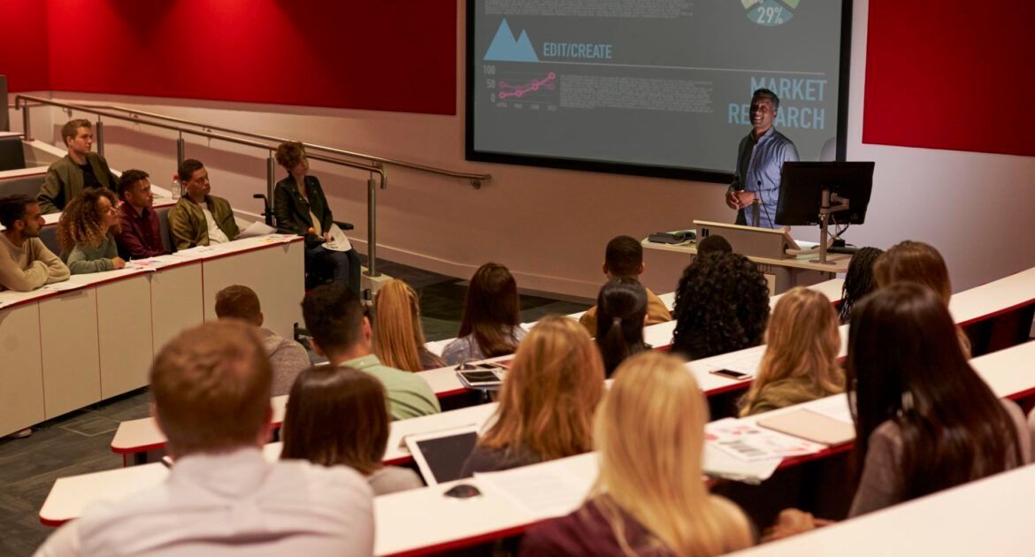 A presenter stands before a lecture hall screen, addressing a group of students seated in tiered rows.