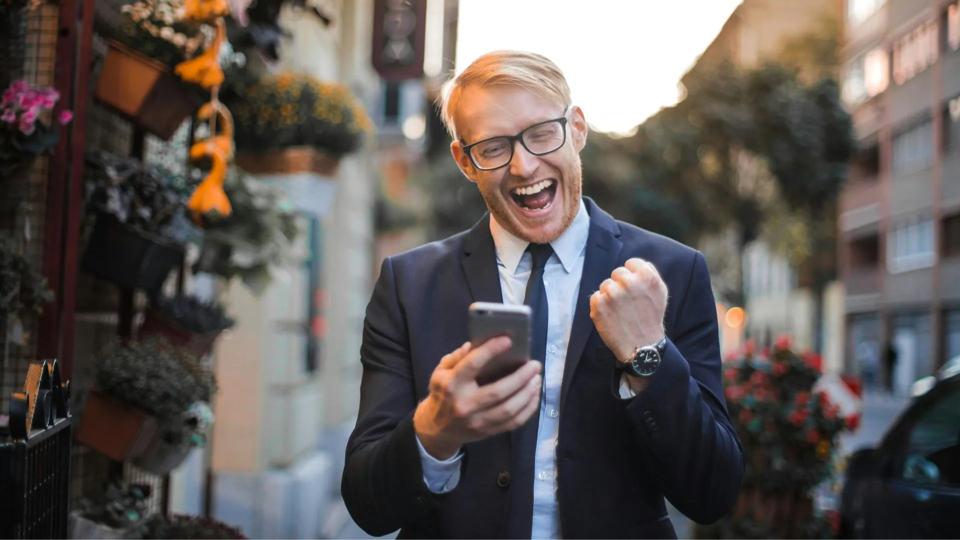 A person in a suit and tie smiling excitedly while holding a smartphone and pumping their fist outdoors.