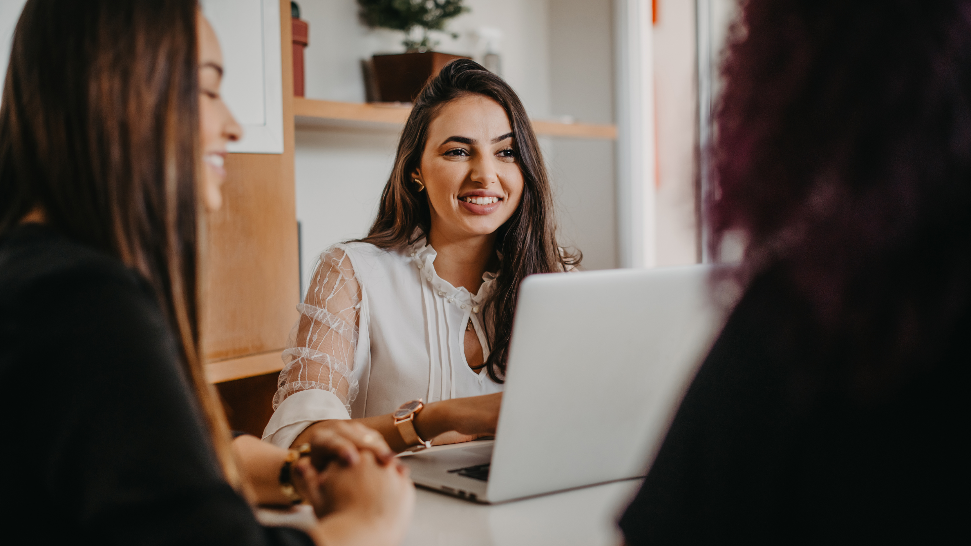 Three people collaborate around a laptop in a brightly lit office setting, focused on a shared work task.