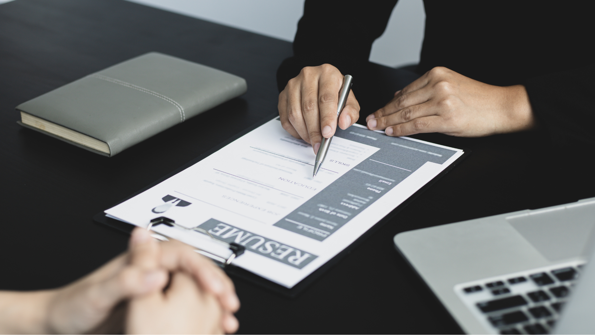 A professional reviews a resume on a black desk with a laptop and notebook nearby.